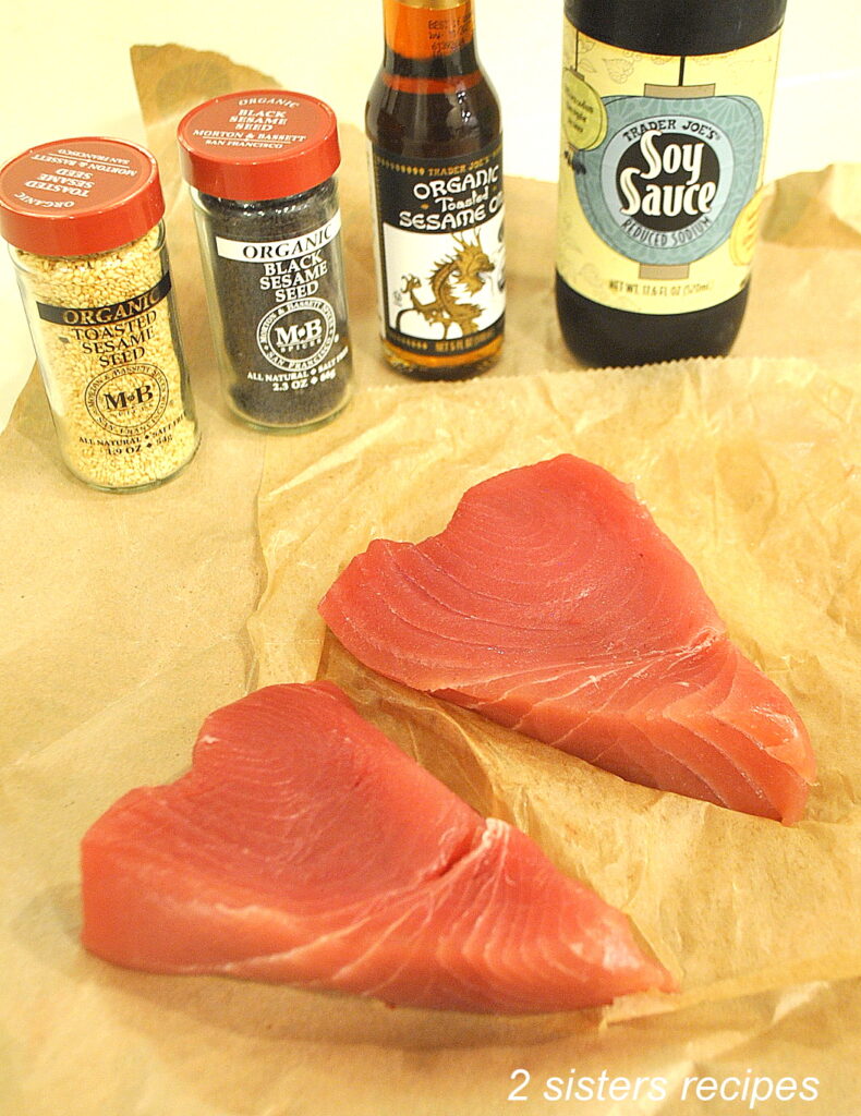Two raw tuna steaks on parchment paper along with bottled soy sauce along with sesame seed oil and jars of sesame seeds.