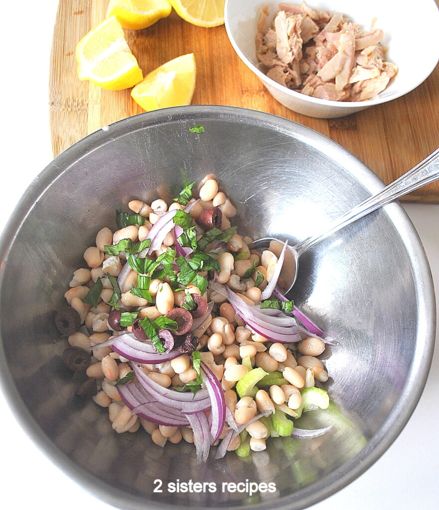 A silver mixing bowl filled with white beans, sliced celery, red onions, capers, and olives.