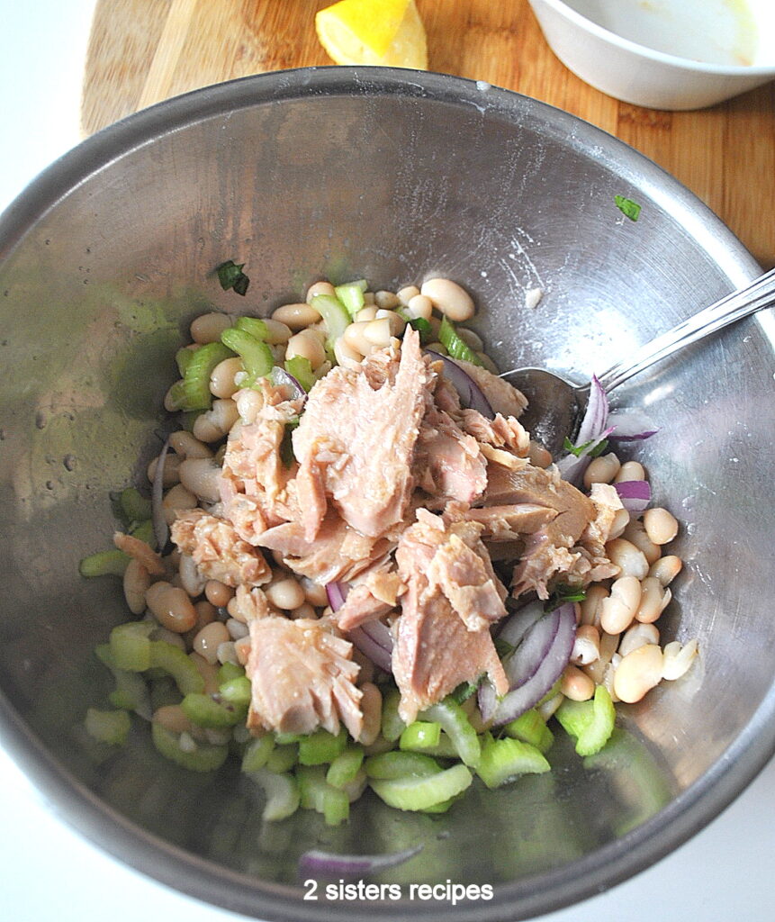 A silver mixing bowl with tuna on top on white beans, celery and capers.
