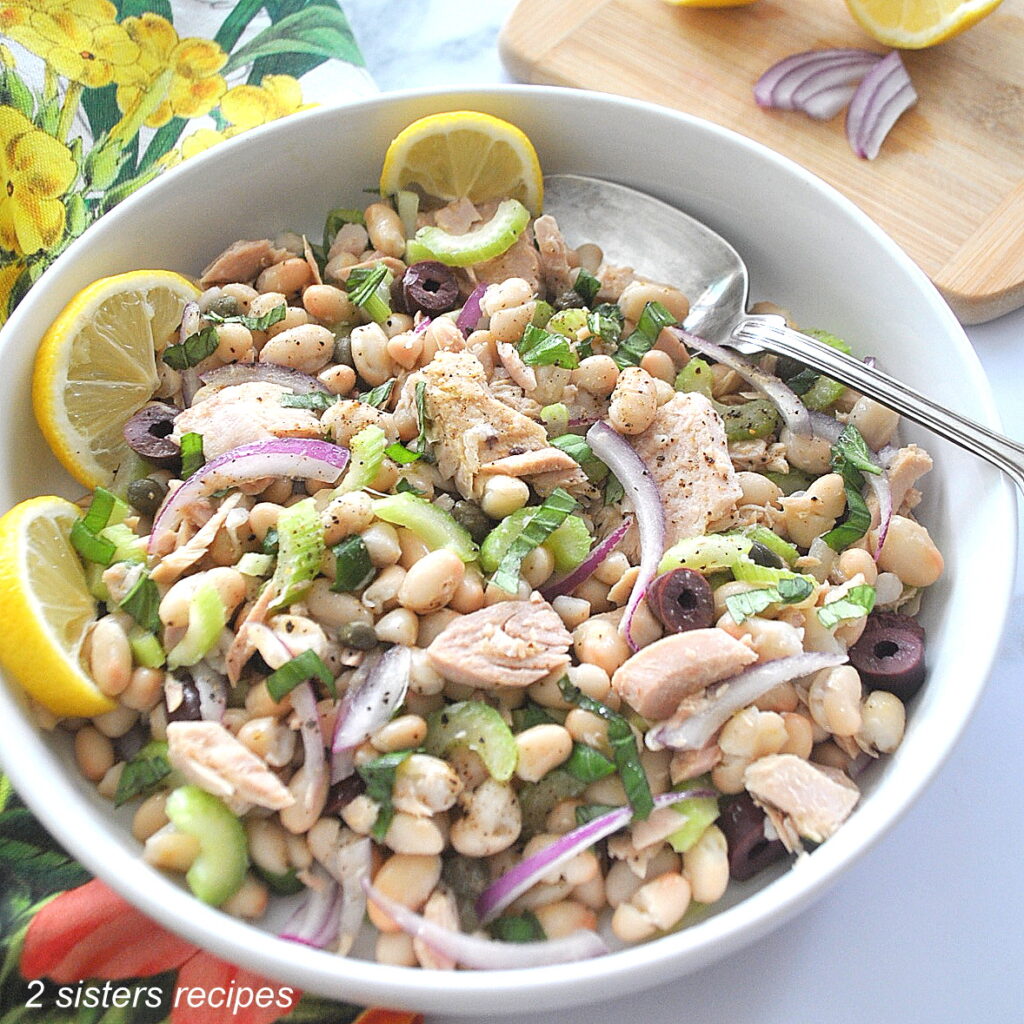 A white bowl filled with a mixture of white beans, tuna, sliced celery and catpers.