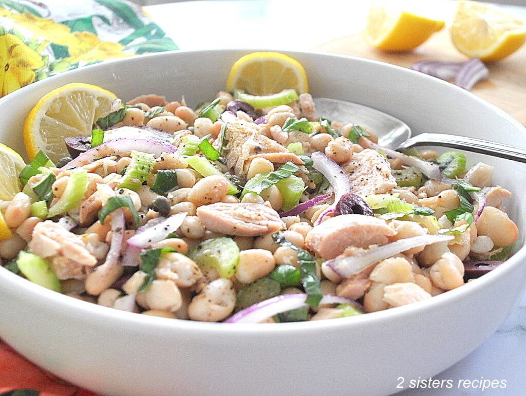 A white bowl filled with a white bean and tuna, celery, and capers salad with lemon wedges.