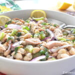 A white bowl filled with a white bean and tuna, celery, and capers salad with lemon wedges.