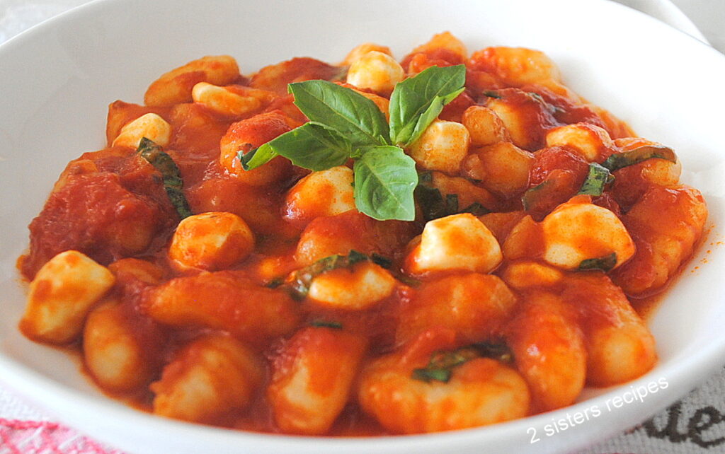 A white dish filled with gnocchi pasta smothered in tomato sauce with pearl mozzarella and basil.