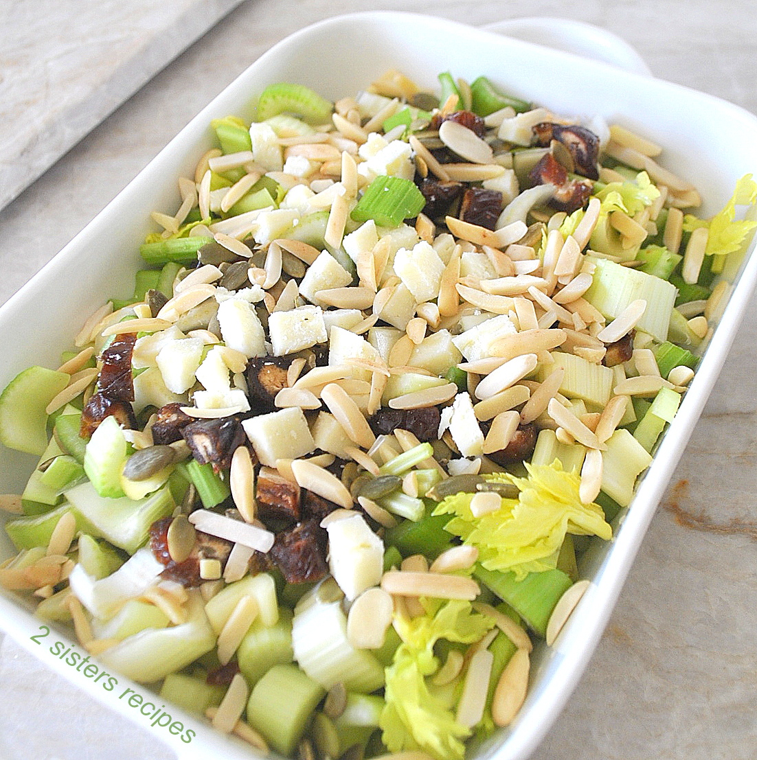 A white dish filled with crunchy celery salad topped with slivered almonds, and chopped pecorino cheese.