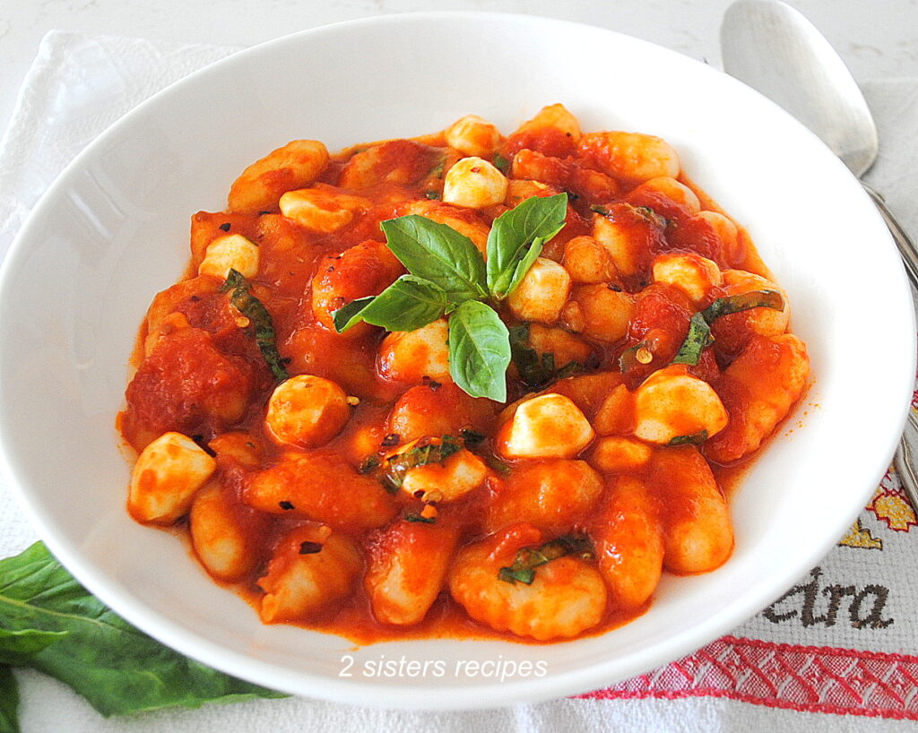 A white dish filled with cooked gnocchi smothered in tomato sauce with mini mozzarella balls and basil.
