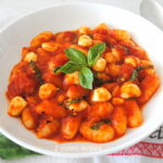 A white dish filled with cooked gnocchi smothered in tomato sauce with mini mozzarella balls and basil.