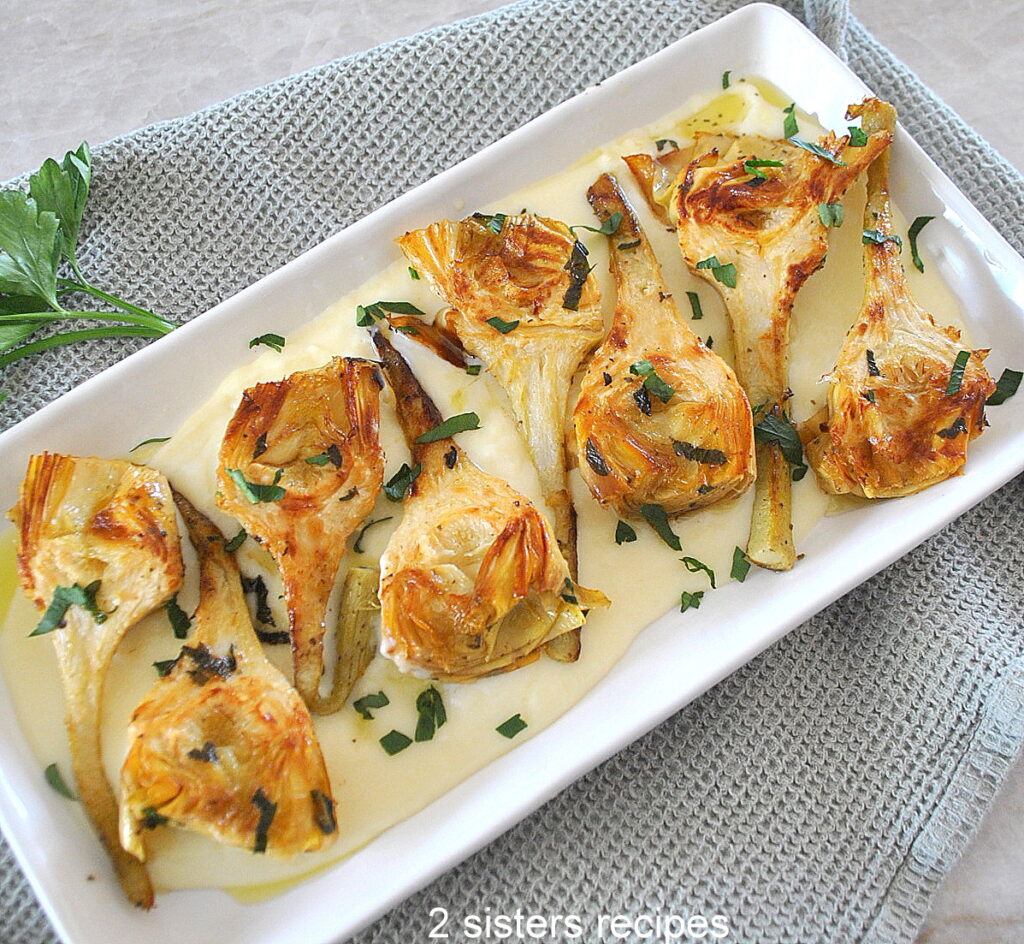 A rectangular dish with a serving of seared artichokes with stems over a cheese cream.
