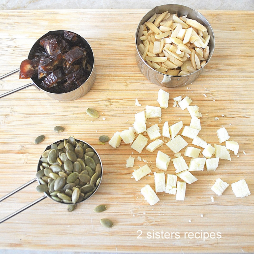 A cutting board with measuring cups with slivered almonds, chopped dates and pumpkin seeds with cubes of cheese.